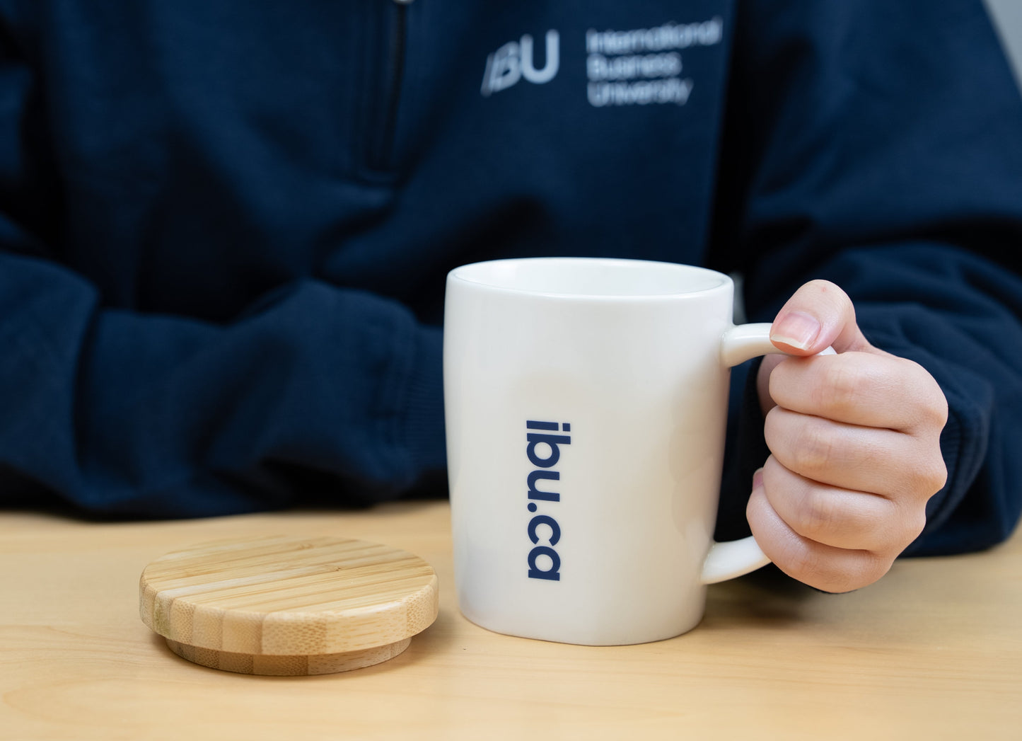 A close-up of the white IBU university mug with the ibu.ca logo, as a student sets the bamboo coaster beside it.