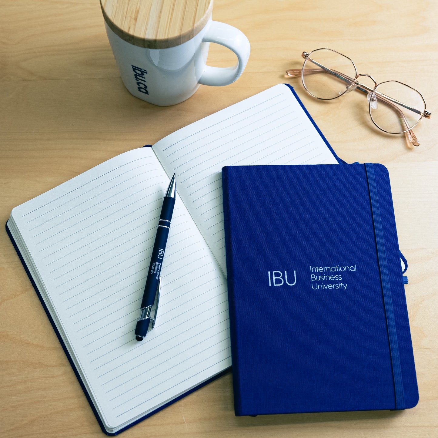 International Business University Notebook on a desk with a pen, mug and glasses from the IBU merch